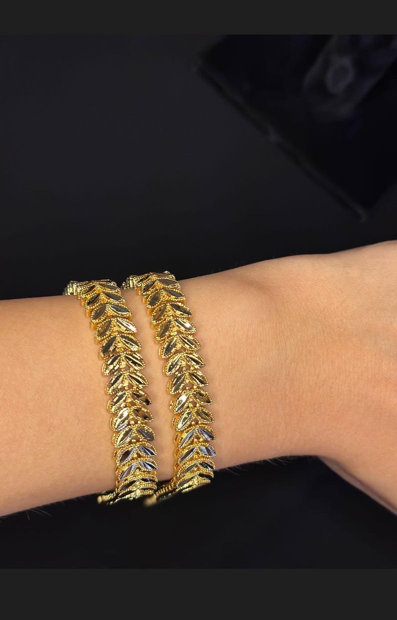 May include: Two gold leaf-shaped bangles on a wrist. The bangles are made of two-toned gold, with a lighter gold color on the inside of the leaves and a darker gold color on the outside.