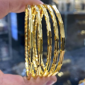 May include: A set of six gold bangles with a faceted design. The bangles are held in a hand, showcasing their bright, reflective surface. The jewelry is likely made of gold and is suitable for everyday wear or special occasions.
