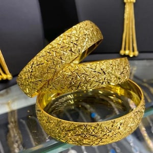 May include: Three gold bangles featuring a textured, cross-hatched pattern across the outer surface. The bracelets are circular with a polished interior finish, reflecting light across the metallic surface to highlight the intricate, repeating geometric design.
