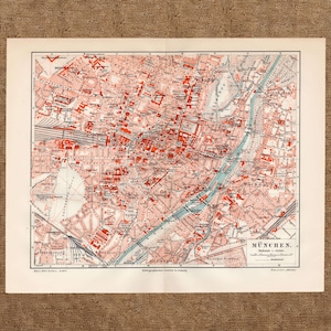 May include: A vintage paper map of Munich, Germany, featuring a grid system with letters and numbers. The map displays streets, buildings in red, and the Isar River in blue. The text MÜNCHEN and a scale are printed in the bottom right corner.