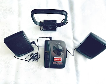 May include: A vintage black Sony Walkman cassette player with attached speakers and headphones. The Walkman has a red and white "Fortuna" sticker. The speakers and headphones are also black. The setup is on a white, patterned surface.