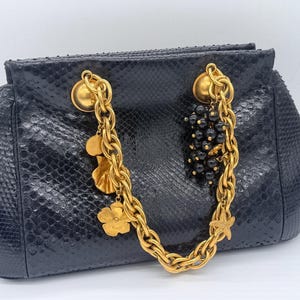 Può includere: Una borsa a mano nera con una trama testurizzata simile alla pelle di serpente, dotata di un manico a catena color oro spesso. Il manico è decorato con ciondoli dorati pendenti, tra cui un fiore e una stella marina, e un grappolo di perline nere.