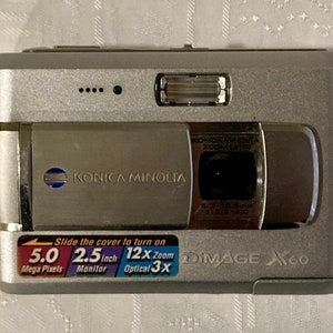 May include: Silver Konica Minolta Dimage X60 digital camera. Features include a 5.0 megapixel sensor, 2.5-inch monitor, and 12x zoom. The camera has a retractable lens and a built-in flash.