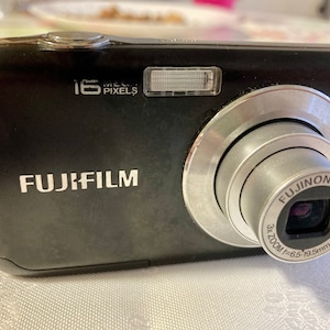 Puede incluir: Cámara digital Fujifilm negra con lente plateada y el texto "16 Mega Pixels". La lente tiene el texto "Fujinon Lens" y "3x Zoom". La cámara está sobre una superficie blanca.