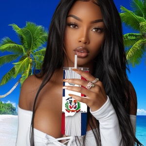 Puede incluir: Una mujer con cabello negro largo sostiene un vaso blanco con la bandera de la República Dominicana impresa en él. Lleva una blusa blanca sin hombros y mira a la cámara.