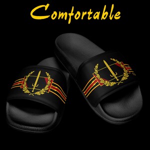 Puede incluir: Sandalias deslizantes negras con un emblema dorado y rojo que presenta una espada, una corona de laurel y rayas. La palabra "Comfortable" está escrita en amarillo encima de las sandalias. Las sandalias están sobre un fondo negro.