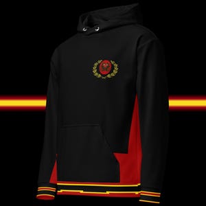 May include: Black hoodie with red side panels and a front pocket. The hoodie features a crest design on the chest and stripes on the cuffs and hem. The design includes gold and red accents.