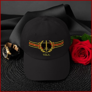 Puede incluir: Gorra de béisbol negra con un emblema dorado y rojo que presenta una espada, una corona de laurel y las letras "T.S.A.". También se ve un reloj de pulsera dorado con una correa de cuero marrón y una rosa roja.