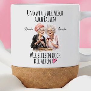 May include: A white ceramic mug with a cork base featuring an illustration of two elderly women holding wine glasses. The text reads: Und wirft der Arsch auch Falten, Klaudia, Kornelia, Wir bleiben doch die Alten, with a small pink heart icon.