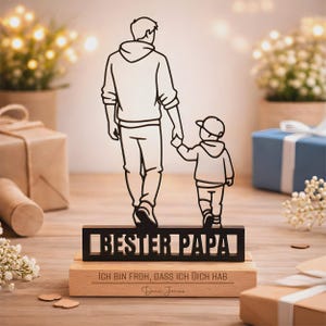 May include: A black metal silhouette of an adult and a child holding hands, mounted on a rectangular wooden base. The metal piece features the text "BESTER PAPA" and the wooden base is engraved with "ICH BIN FROH, DASS ICH DICH HAB" and "Dein Jonas".