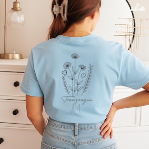 以下が含まれることがあります： 背中に黒い花の模様が入った水色のTシャツ。デザインには3つの花、ハート、筆記体で「Trauzeugin」という言葉が含まれています。シャツは柔らかい素材でできており、半袖です。