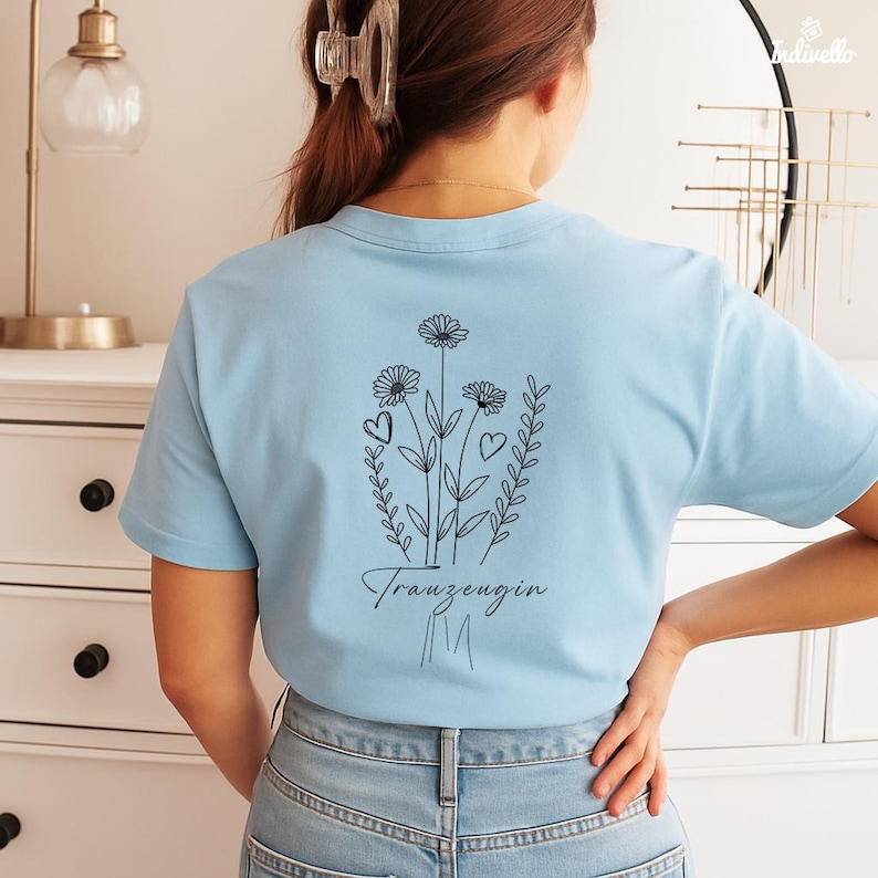 以下が含まれることがあります： 背中に黒い花の模様が入った水色のTシャツ。デザインには3つの花、ハート、筆記体で「Trauzeugin」という言葉が含まれています。シャツは柔らかい素材でできており、半袖です。