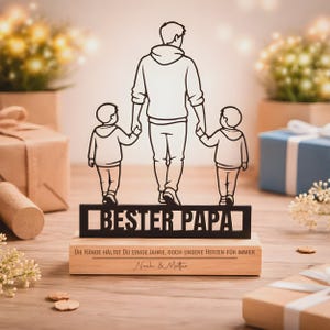 May include: A black metal silhouette of an adult holding the hands of two children, mounted on a rectangular wooden base. The metal cutout features the text "BESTER PAPA" and the wooden base is engraved with "DIE H&Auml;NDE H&Auml;LTST DU EINIGE JAHRE, DOCH UNSERE HERZEN F&Uuml;R IMMER" and "Noah & Matteo".