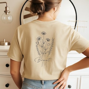 以下が含まれることがあります： 背中に花の模様が入ったベージュ色のTシャツ。デザインには3つの花、ハート、筆記体で「Braut」の文字が含まれています。柔らかい素材でできており、半袖です。