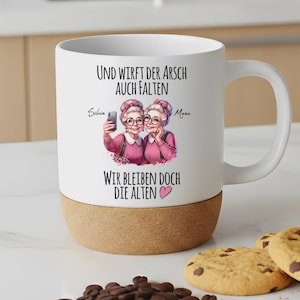 May include: A white ceramic mug with a cork base featuring a graphic of two older individuals with glasses and pink sweaters taking a selfie. The text reads: Und wirft der Arsch auch Falten, wir bleiben doch die Alten, with the names Silvia and Maru.