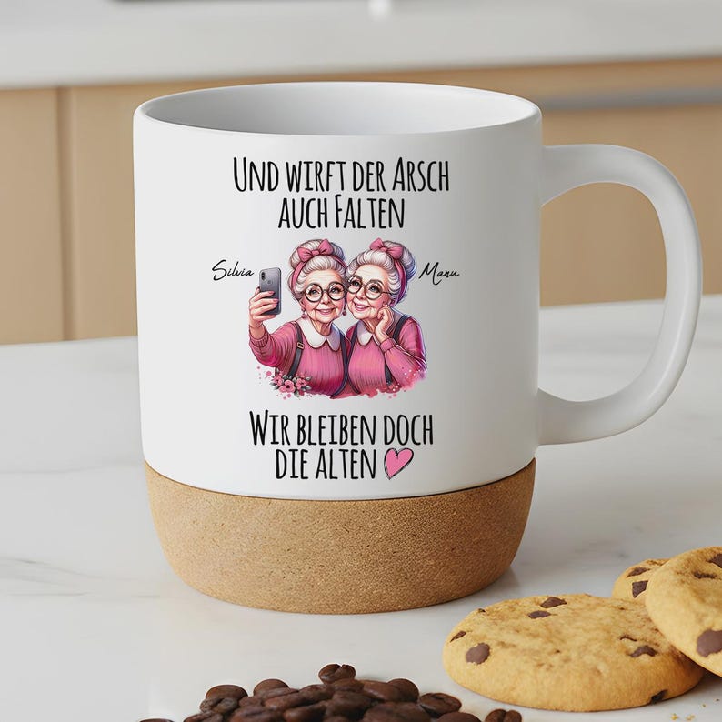 May include: A white ceramic mug with a cork base featuring a graphic of two older individuals with glasses and pink sweaters taking a selfie. The text reads: Und wirft der Arsch auch Falten, wir bleiben doch die Alten, with the names Silvia and Maru.