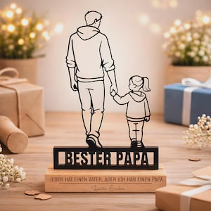 May include: A black metal silhouette of an adult and a child holding hands, mounted on a rectangular wooden base. The metal piece features the text "BESTER PAPA" and the wooden base is engraved with "JEDER HAT EINEN VATER, ABER ICH HAB EINEN PAPA" and "Deine Emilia".