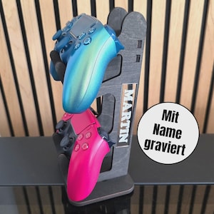 Könnte beinhalten: Ein grauer Holz-Gaming-Controller-Ständer, der zwei Controller hält, einen blauen und einen pinkfarbenen. Der Ständer hat den Namen "MARTIN" eingraviert. Ein weißer Kreis mit dem Text "Mit Name graviert" ist ebenfalls sichtbar.