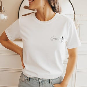 以下が含まれることがあります： 白いTシャツに筆記体の「Jessica」という名前と小さな花の模様がプリントされています。Tシャツはクルーネックで、柔らかい素材でできているようです。デザインは左胸にプリントされています。