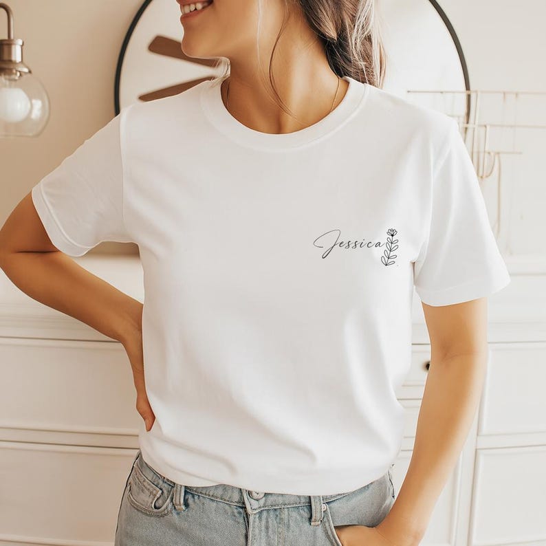 以下が含まれることがあります： 白いTシャツに筆記体の「Jessica」という名前と小さな花の模様がプリントされています。Tシャツはクルーネックで、柔らかい素材でできているようです。デザインは左胸にプリントされています。