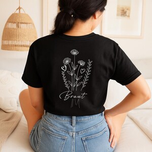 以下が含まれることがあります： 背中に白い花の模様が入った黒いTシャツ。デイジー、ハート、筆記体で「Braut」の文字が入っています。シャツはブルージーンズと組み合わせています。