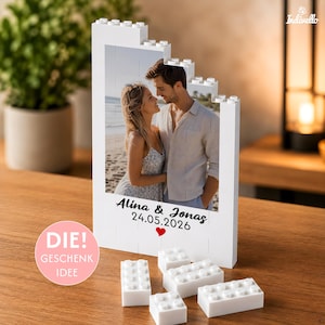 Könnte beinhalten: Ein weißer Bausteinrahmen mit einem Foto eines Paares am Strand. Der Rahmen enthält den aufgedruckten Text Alina & Jonas 24.05.2026 mit einem kleinen roten Herz. Vor dem Rahmen liegen mehrere lose weiße Bausteine auf der Oberfläche.