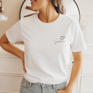 以下が含まれることがあります： クルーネックの白いTシャツ。Tシャツには、筆記体で「Jessica」という名前と小さなハートのシンボルが描かれています。このシャツは柔らかい素材でできており、カジュアルウェアに適しています。