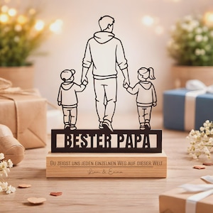 May include: A black metal silhouette of a parent holding hands with two children, standing on a rectangular wooden base. The metal cutout features the text BESTER PAPA. The wooden base is engraved with the text DU ZEIGST UNS JEDEN EINZELNEN WEG AUF DIESER WELT and the names Liam & Emma.