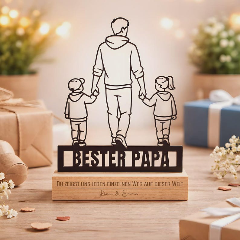 May include: A black metal silhouette of a parent holding hands with two children, standing on a rectangular wooden base. The metal cutout features the text BESTER PAPA. The wooden base is engraved with the text DU ZEIGST UNS JEDEN EINZELNEN WEG AUF DIESER WELT and the names Liam & Emma.