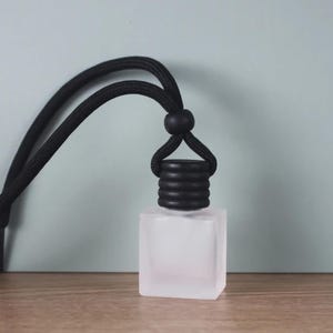 May include: A frosted glass, square-shaped air freshener bottle with a black cap and a black cord for hanging. The bottle sits on a wooden surface against a light teal background.