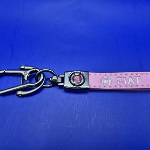May include: A pink leather key ring with a silver metal loop and a silver metal charm featuring the Fiat logo. The key ring has the word "FIAT" embossed on it.
