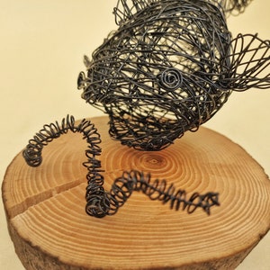 Op de afbeelding: Een draadbeeld van een vis met een gekrulde staart op een houten voet. De vis is gemaakt van zwart draad en heeft een gedetailleerd, realistisch ontwerp.