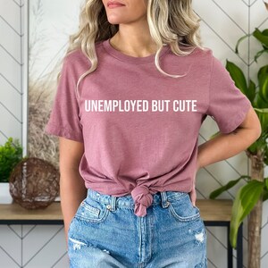 May include: A woman wearing a mauve t-shirt with the text "Unemployed But Cute" in white lettering. She is wearing denim shorts and has her hair pulled back.