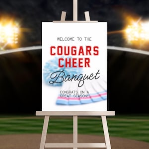 May include: A white sign on a wooden easel with text that reads "Welcome to the Cougars Cheer Banquet." The text is in red and black. A light blue pom-pom and cheerleading skirt are in the background. The sign also says "Congrats on a great season!"