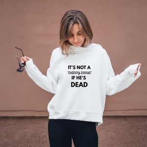 May include: White hoodie with the text "IT'S NOT A 'daddy issue' IF HE'S DEAD" in black. The hoodie is worn by a person holding glasses.