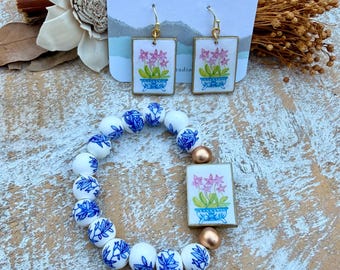 May include: A set of jewelry featuring rectangular earrings and a beaded bracelet. The earrings and the bracelet centerpiece display a painted pink flower in a blue pot. The bracelet includes white beads with blue floral patterns and two gold beads.