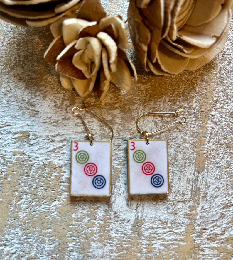 Op de afbeelding: Een paar goudkleurige oorbellen. Elke oorbel heeft een rechthoekige witte tegel met het nummer "3" en drie kleurrijke cirkelvormige ontwerpen: groen, rood en blauw. De oorbellen worden gepresenteerd op een licht houten oppervlak.