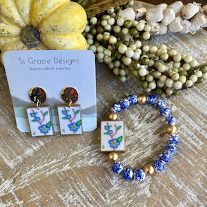 K&ouml;nnte beinhalten: Handgefertigtes Schmuckset mit Ohrringen und einem Armband. Die Ohrringe haben ein rechteckiges Design mit floralem Muster und goldenen Akzenten. Das Armband hat blaue und wei&szlig;e Perlen mit goldenen Akzenten.