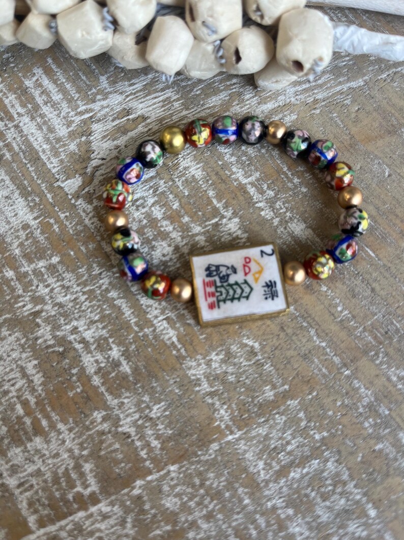 Op de afbeelding: Een kralenarmband met kleurrijke, patroon kralen en goudkleurige tussenkralen. De armband bevat een rechthoekige bedel met Chinese karakters en het nummer 2.
