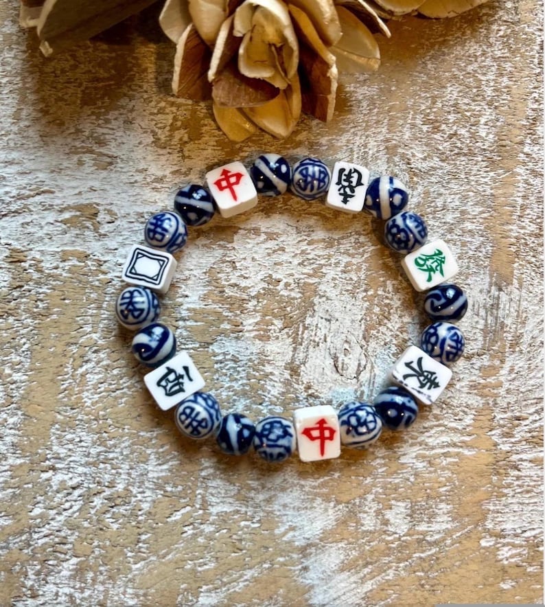 May include: A beaded bracelet featuring a mix of white square beads with Chinese characters and blue and white patterned round beads. The bracelet is displayed on a light-colored, textured surface.