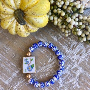 May include: A stretch bracelet featuring round beads with a blue and white floral pattern, two smooth gold-toned spherical beads, and a rectangular white tile charm decorated with a painted blue flower branch and the green number three.