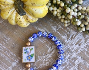 May include: A stretch bracelet featuring round beads with a blue and white floral pattern, two smooth gold-toned spherical beads, and a rectangular white tile charm decorated with a painted blue flower branch and the green number three.