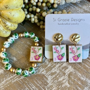 K&ouml;nnte beinhalten: Ein handgefertigtes Schmuckset mit Armband und Ohrringen. Das Armband hat wei&szlig;e und gr&uuml;ne Perlen mit goldenen Akzenten. Die Ohrringe haben goldene Stecker und rechteckige Anh&auml;nger mit floralen Designs und der Zahl 3.