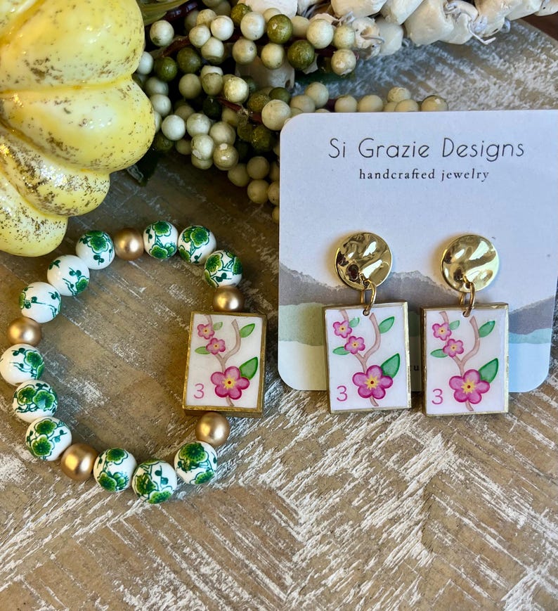 K&ouml;nnte beinhalten: Ein handgefertigtes Schmuckset mit Armband und Ohrringen. Das Armband hat wei&szlig;e und gr&uuml;ne Perlen mit goldenen Akzenten. Die Ohrringe haben goldene Stecker und rechteckige Anh&auml;nger mit floralen Designs und der Zahl 3.