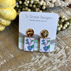 K&ouml;nnte beinhalten: Handgefertigte Ohrringe mit goldfarbenen Oberteilen und rechteckigen Anh&auml;ngern. Die Anh&auml;nger zeigen ein Blumenmuster mit blauen Blumen, gr&uuml;nen Bl&auml;ttern und der Zahl 4. Die Ohrringe befinden sich auf einer wei&szlig;en Karte mit dem Text "Si Grazie Designs".