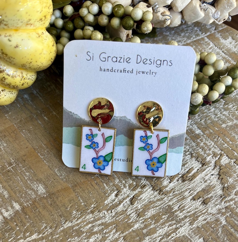 K&ouml;nnte beinhalten: Handgefertigte Ohrringe mit goldfarbenen Oberteilen und rechteckigen Anh&auml;ngern. Die Anh&auml;nger zeigen ein Blumenmuster mit blauen Blumen, gr&uuml;nen Bl&auml;ttern und der Zahl 4. Die Ohrringe befinden sich auf einer wei&szlig;en Karte mit dem Text "Si Grazie Designs".