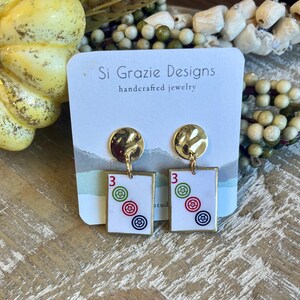 May include: Gold-tone stud earrings with a textured, circular top and a rectangular pendant. The pendant features the number "3" and three concentric circles in red, green, and blue. The earrings are from "Si Grazie Designs, handcrafted jewelry."
