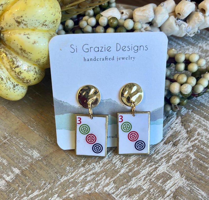 May include: Gold-tone stud earrings with a textured, circular top and a rectangular pendant. The pendant features the number "3" and three concentric circles in red, green, and blue. The earrings are from "Si Grazie Designs, handcrafted jewelry."
