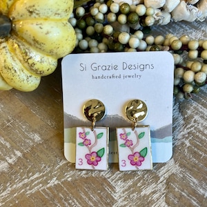 K&ouml;nnte beinhalten: Handgefertigte Ohrringe mit goldfarbenen, geh&auml;mmerten Scheibenaufs&auml;tzen und rechteckigen Anh&auml;ngern. Die Anh&auml;nger zeigen ein Blumendesign mit rosa Bl&uuml;ten, gr&uuml;nen Bl&auml;ttern und der Zahl 3. Die Ohrringe sind von Si Grazie Designs.