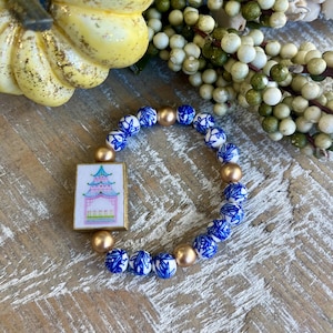 K&ouml;nnte beinhalten: Ein Armband mit blauen und wei&szlig;en Porzellanperlen und goldfarbenen Perlen. Es hat einen rechteckigen Anh&auml;nger mit einem Pagoden-Design in Rosa, Gr&uuml;n und Blau. Das Armband wird auf einer Holzoberfl&auml;che pr&auml;sentiert.